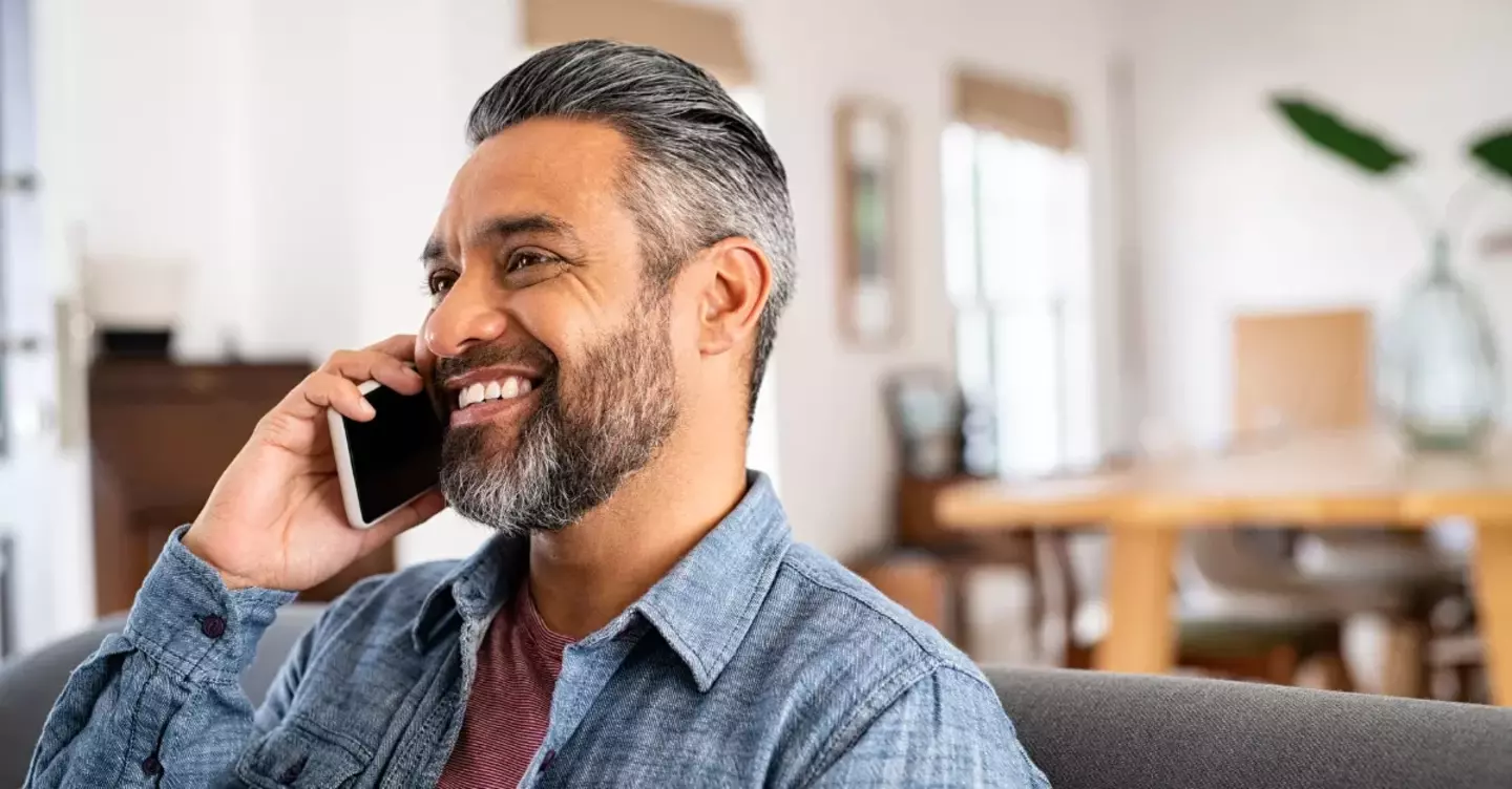 A cheerful man talks on a smartphone, seated comfortably indoors in a bright, casually furnished home.
