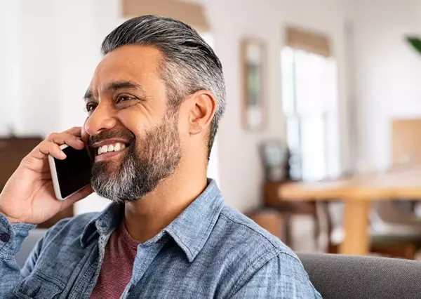 A cheerful man talks on a smartphone, seated comfortably indoors in a bright, casually furnished home.