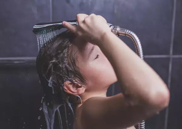 A child holds a showerhead over their head, rinsing shampoo in a tiled shower. Water streams onto hair while eyes are closed, emphasizing a sense of calm cleanliness.