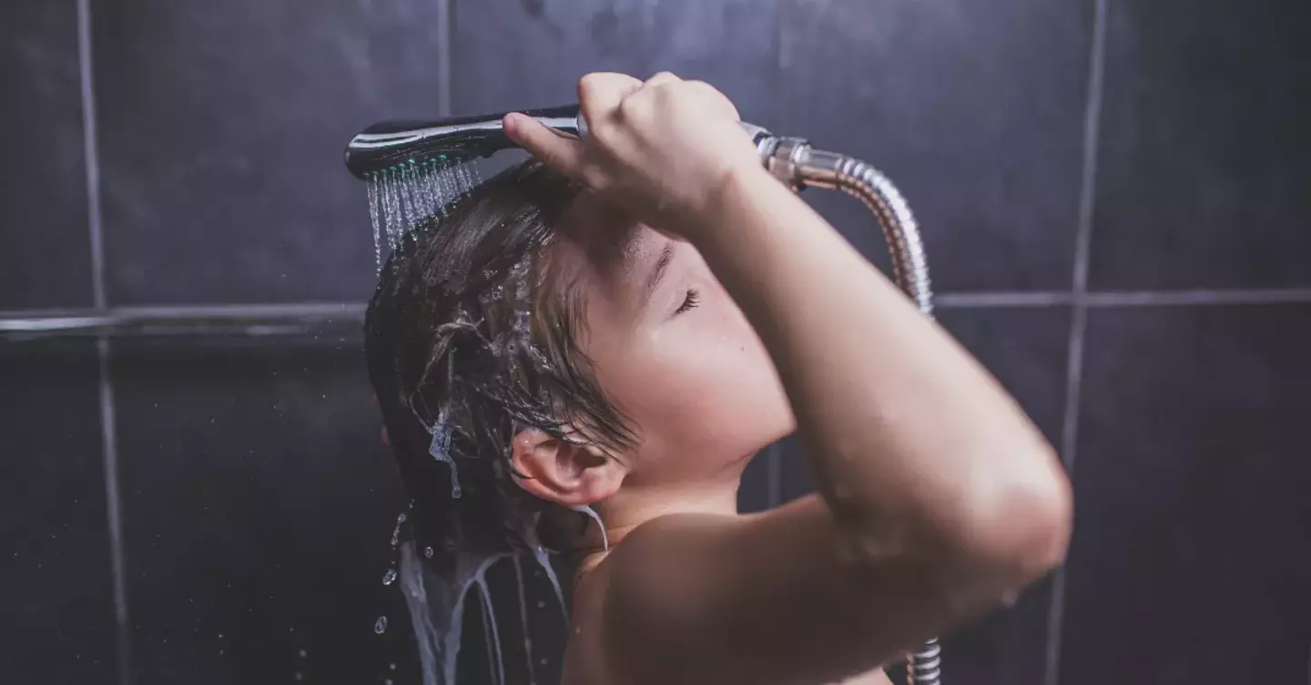 A child holds a showerhead over their head, rinsing shampoo in a tiled shower. Water streams onto hair while eyes are closed, emphasizing a sense of calm cleanliness.