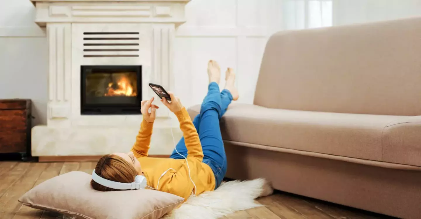 A woman reclines on a pillow, using her smartphone and wearing headphones, in a cozy living room with a lit fireplace.