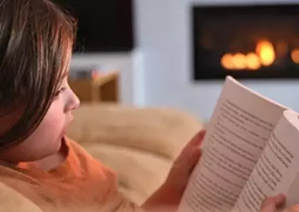 A young girl, wrapped in an orange blanket, reads a book intently, seated by a fireplace that gently illuminates the cozy room.