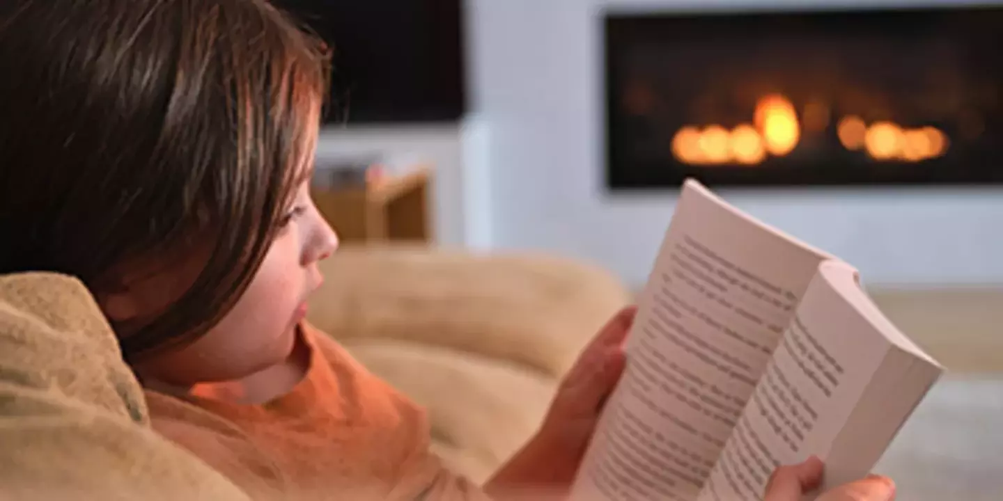 A young girl, wrapped in an orange blanket, reads a book intently, seated by a fireplace that gently illuminates the cozy room.