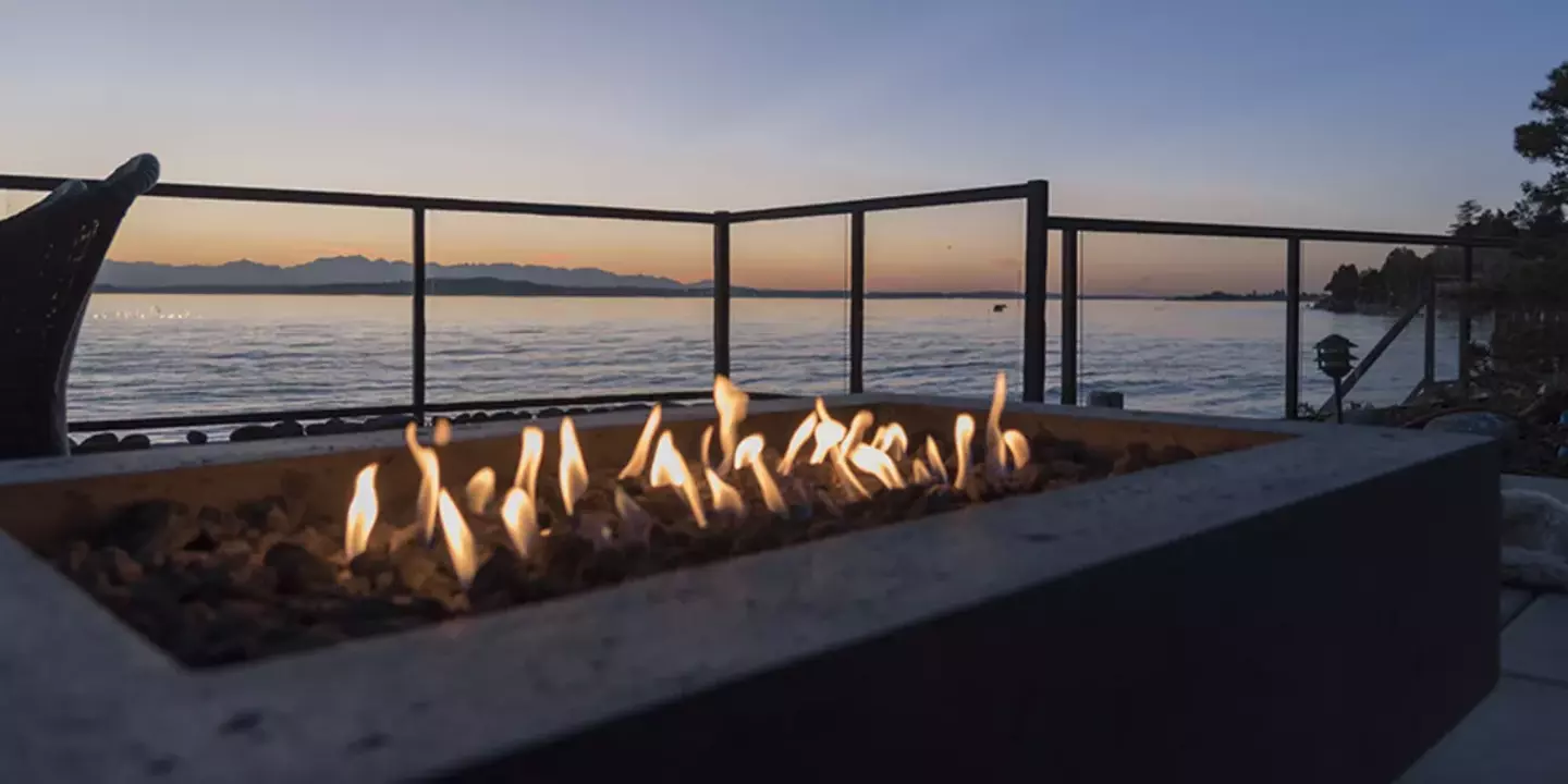 A rectangular fire pit burns warmly on a patio overlooking a calm lake with distant mountains under a dusky sky. A bench faces the serene view, inviting relaxation.