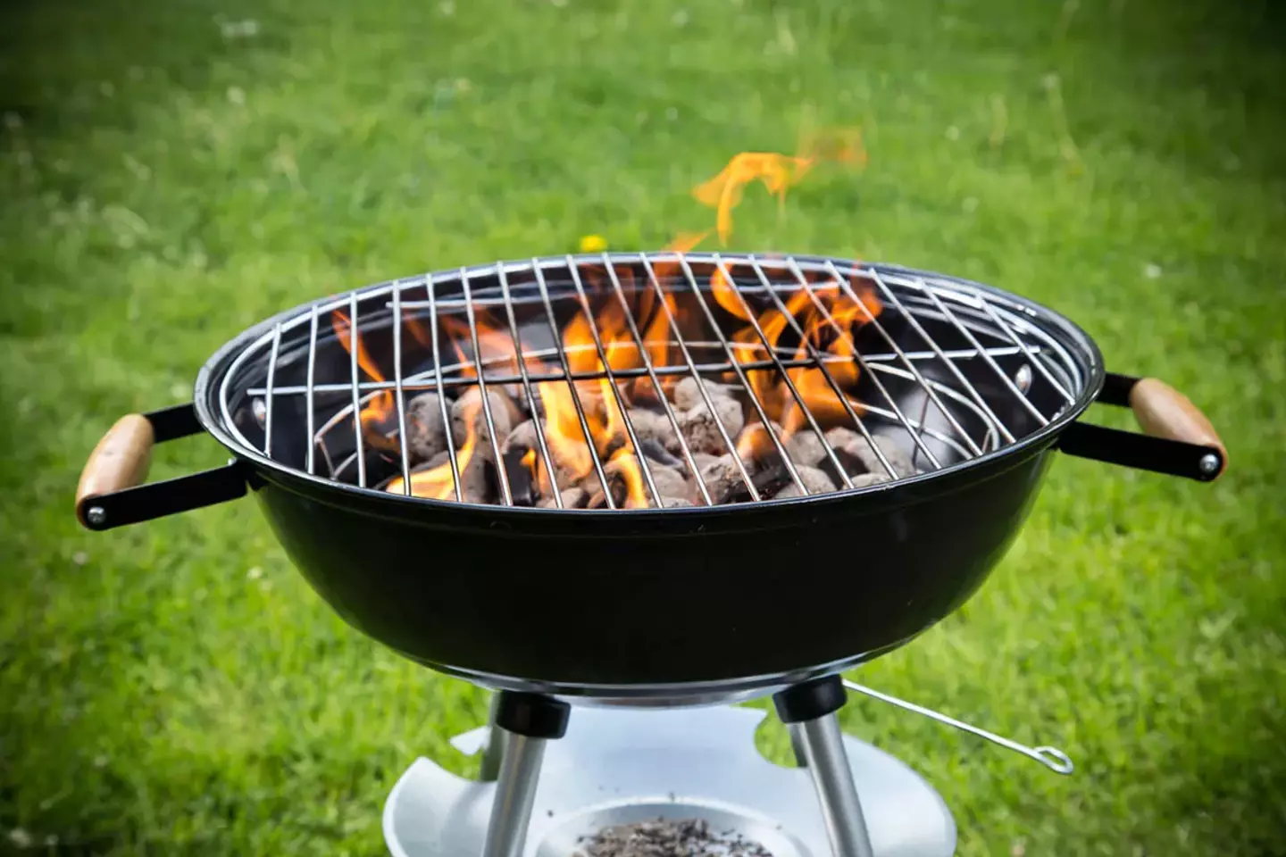A charcoal grill, with flames flickering through a grill grate, stands on vivid green grass, symbolizing outdoor cooking.
