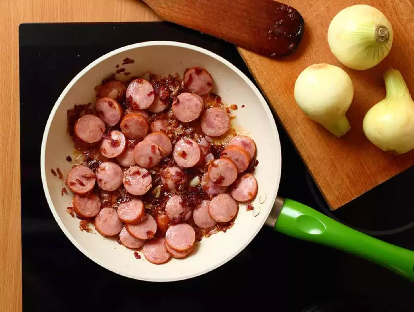 Sliced sausages cook in a pan, caramelizing onions scattered among them. Nearby, whole onions rest on a wooden cutting board, hinting at kitchen meal preparation.