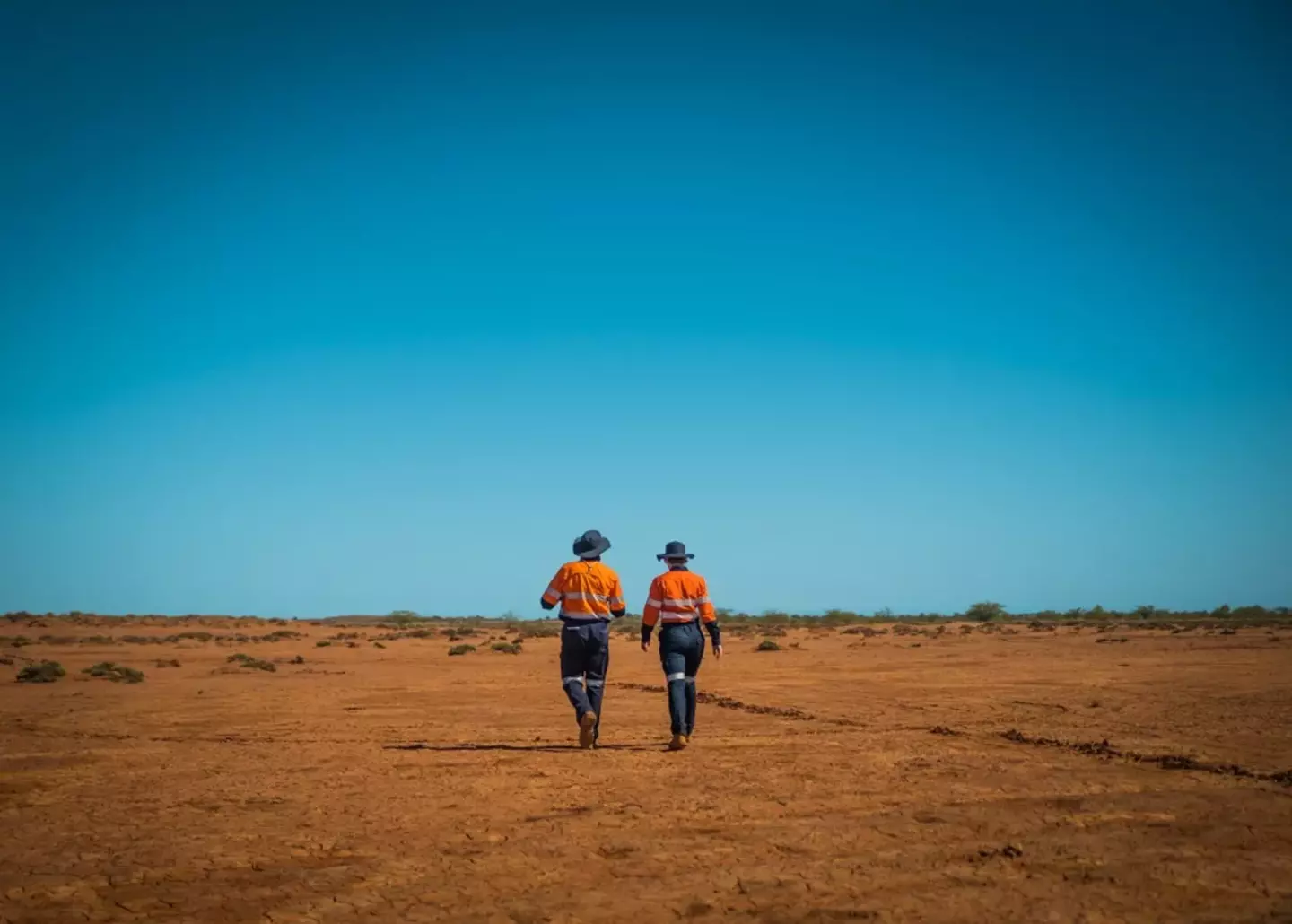 Two workers in orange hi-vis shirts and hats walk away into a vast, barren desert under a clear blue sky.