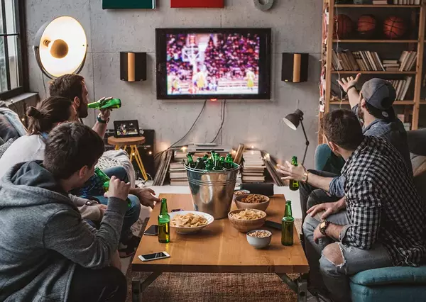 A group of friends watches a basketball game on TV, reacting excitedly in a cozy, well-decorated living room with drinks and snacks.