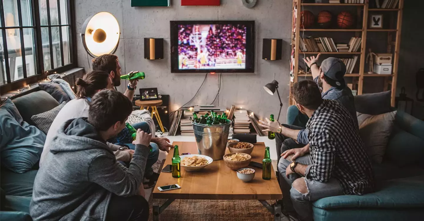 A group of friends watches a basketball game on TV, reacting excitedly in a cozy, well-decorated living room with drinks and snacks.
