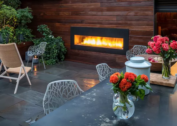 An outdoor patio features a glowing rectangular fireplace set against a wooden wall, surrounded by green foliage, modern furniture, and vibrant flowers, creating a cozy atmosphere.