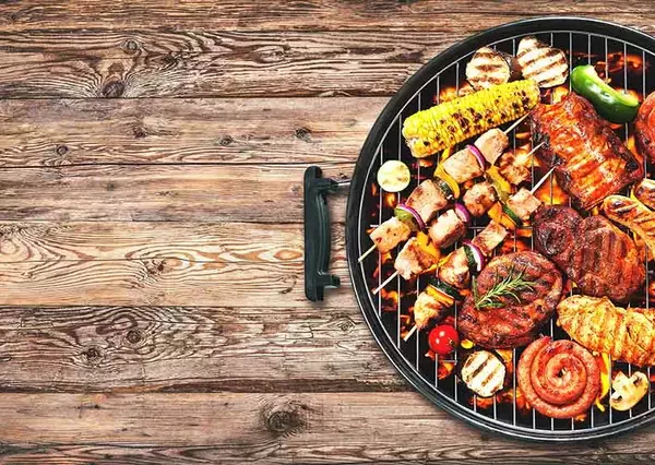 Various meats and vegetables are grilling on a barbecue, emitting steam over glowing coals, set against a rustic wooden background.