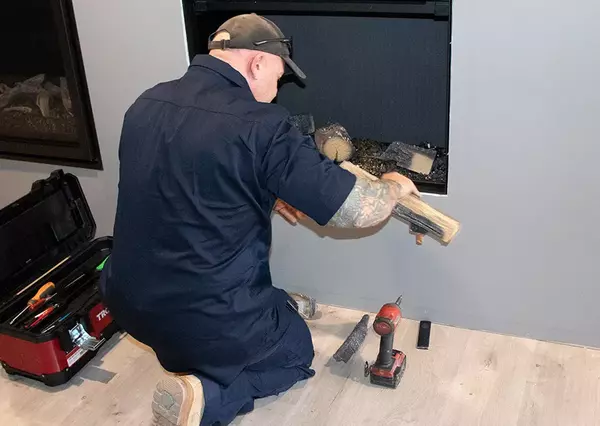 A technician in a dark blue uniform is installing logs into a modern fireplace. He is kneeling beside an open toolbox and a cordless drill, in a well-lit living room.