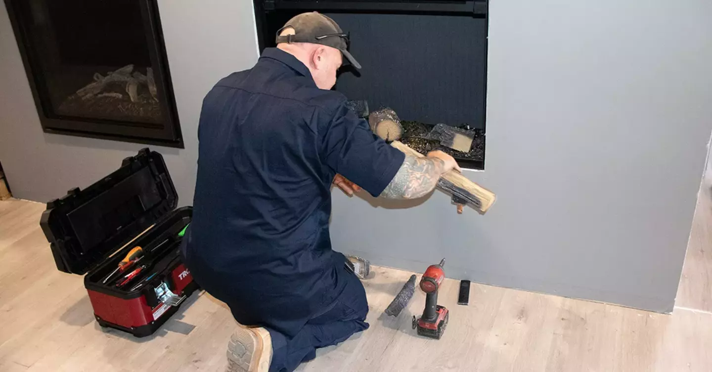 A technician in a dark blue uniform is installing logs into a modern fireplace. He is kneeling beside an open toolbox and a cordless drill, in a well-lit living room.