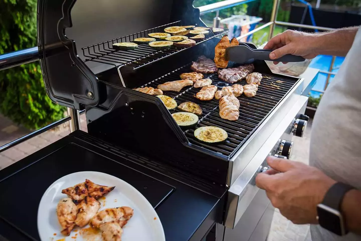 A person grills an assortment of meat and vegetables on a barbecue in a lush backyard, transferring cooked food to a nearby white plate.