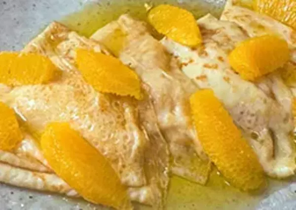 Crepes are garnished with orange slices and drizzled with syrup, served on a textured plate.