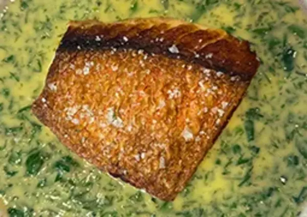 A piece of crispy-skinned salmon rests atop a creamy, herb-flecked sauce served in a textured, beige bowl.