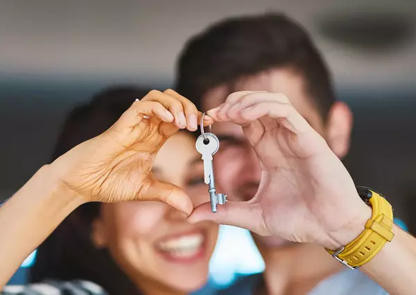A couple forms a heart shape with their hands around a key, smiling joyfully, symbolizing new beginnings or ownership, set against a softly blurred indoor background.