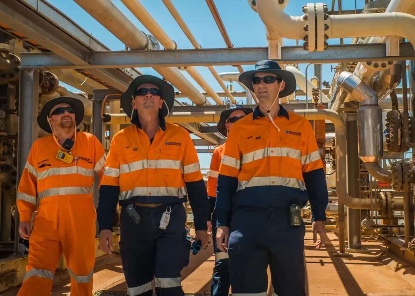 Four workers in high-visibility orange and blue uniforms inspect a complex of metal pipes and valves at an industrial site, conveying a sense of focused oversight and safety.