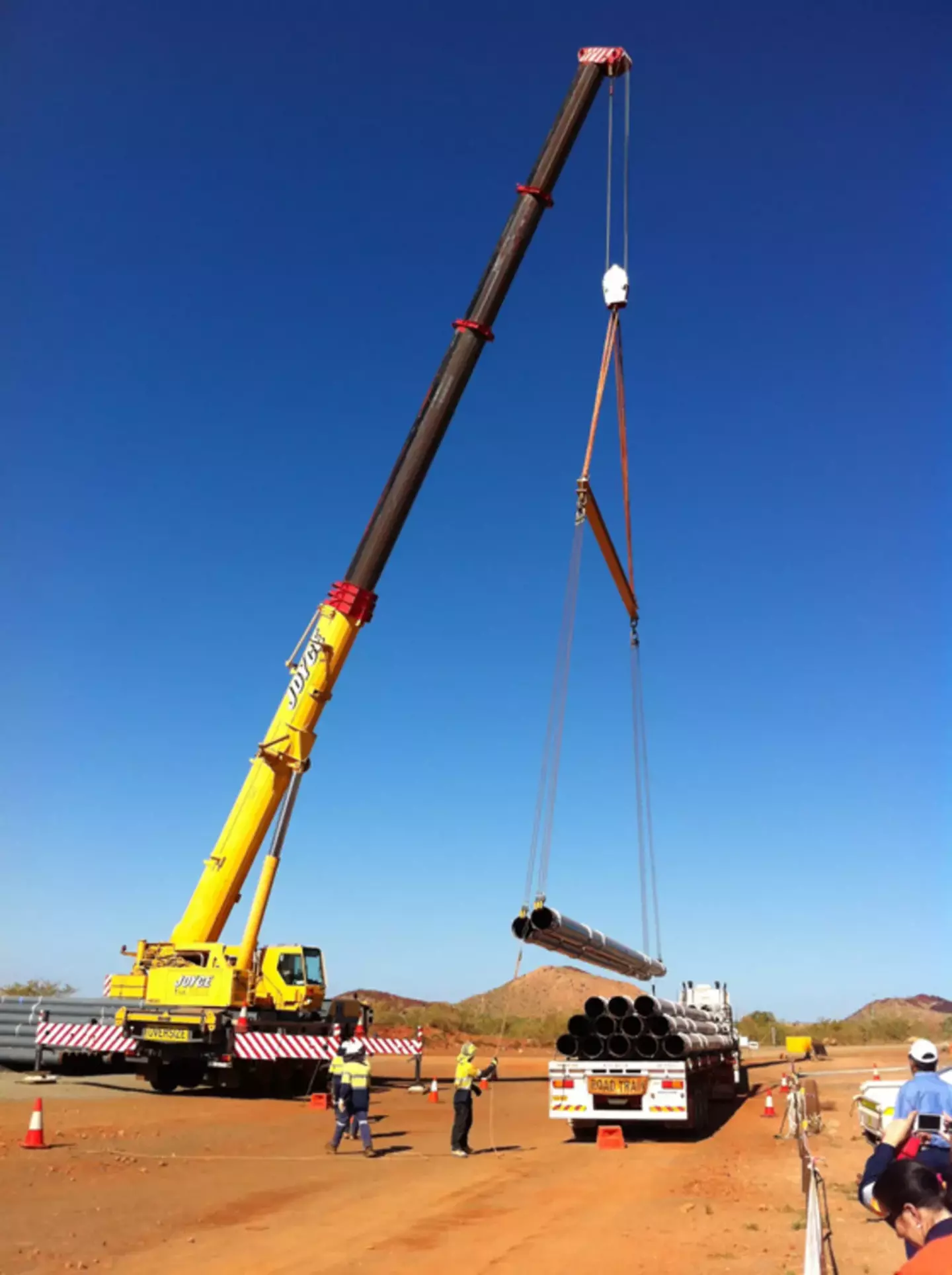 A large yellow crane lifts heavy pipes at a construction site under a clear blue sky, with workers supervising. There is no text in the image.