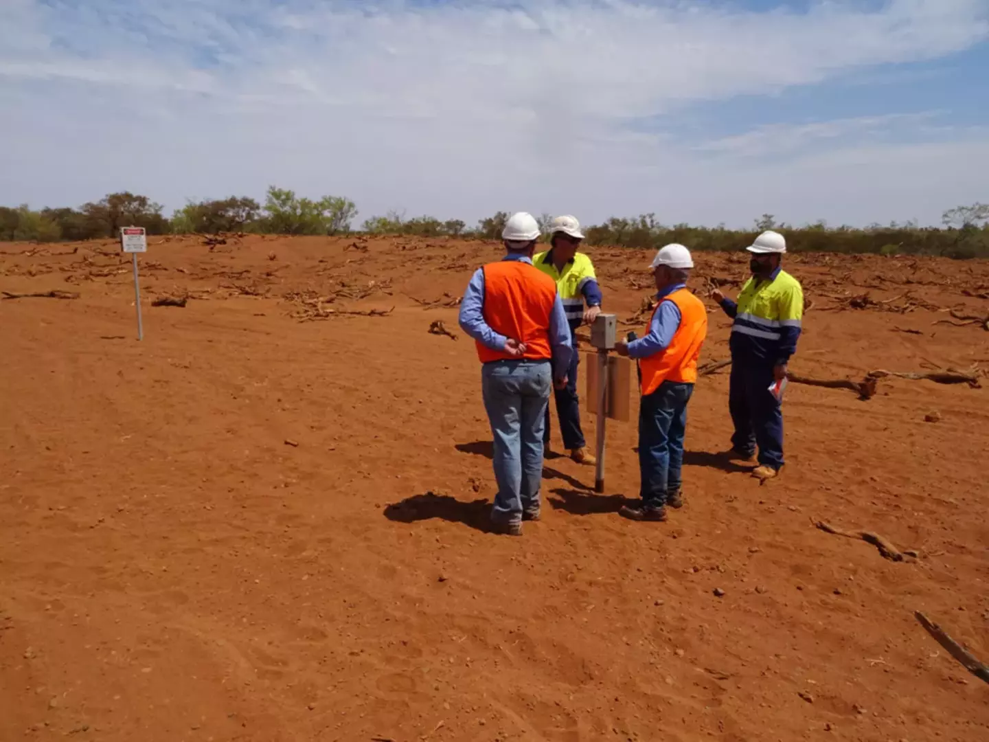 Five construction workers in helmets and high-visibility clothing discuss near a post in a vast, red dirt field under a clear blue sky. A sign reads "DANGER Construction site Keep out."