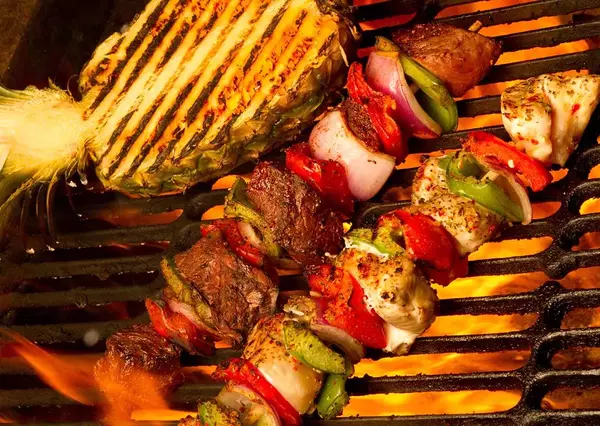 Various foods including corn, meat, and skewered vegetables are grilling over an open flame, showcasing a vibrant display of cooking on a barbecue.