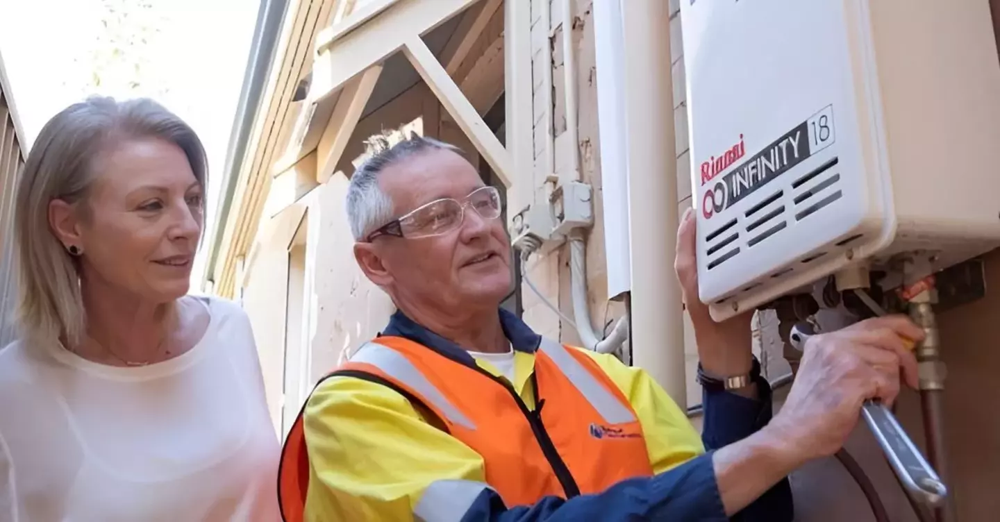 A technician in a high-visibility vest installs a Rinnai Infinity 18 water heater as a homeowner observes, in a narrow outdoor alley by a residential building.