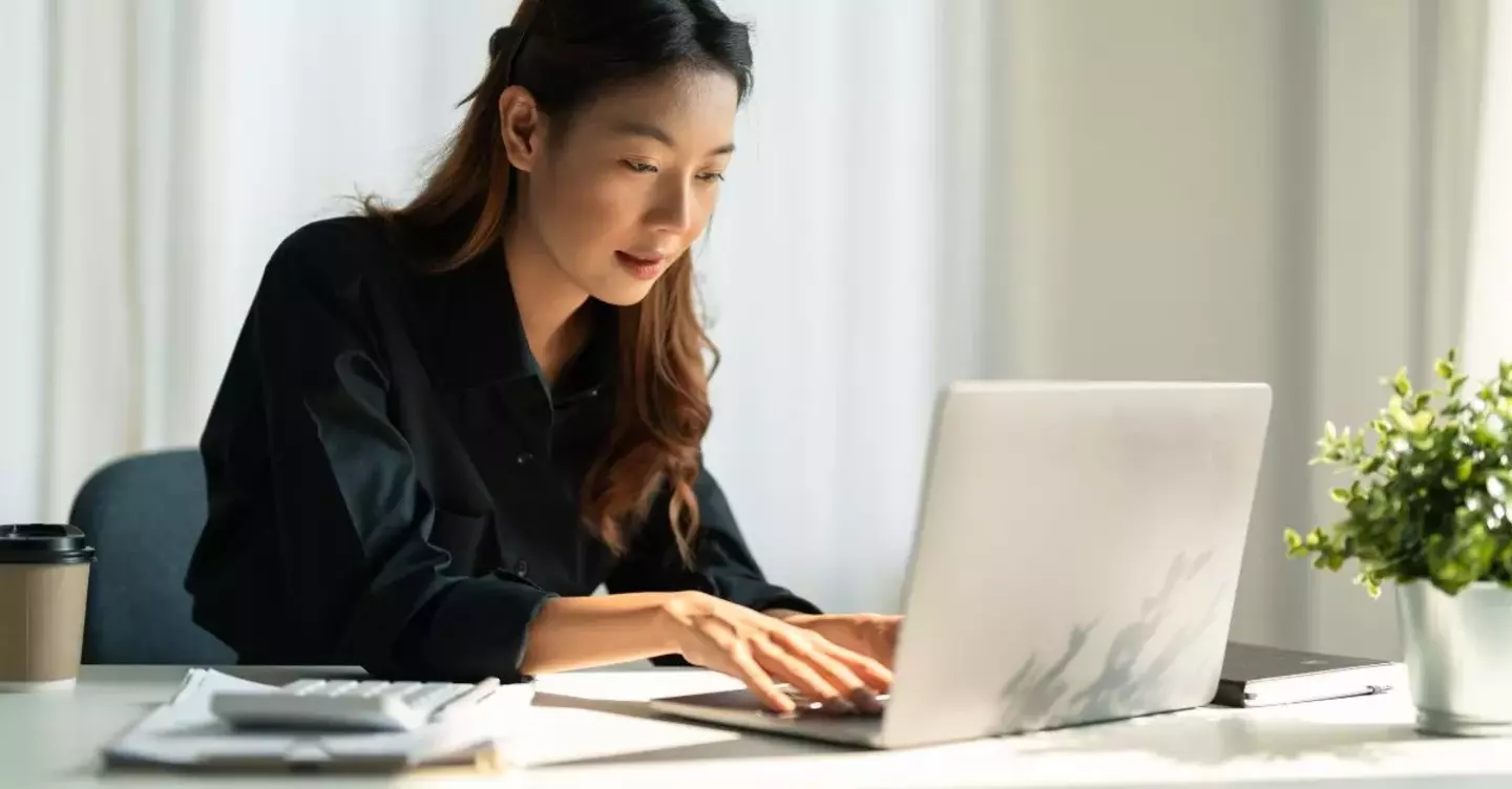 A woman is focused intently on typing on a laptop in a sunlit office with a tidy desk, a coffee cup, and a small plant.