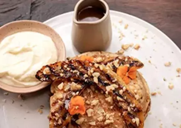 Grilled bananas topped with nuts and syrup served with whipped cream and a side of sauce on a white plate, set on a dark wooden table.