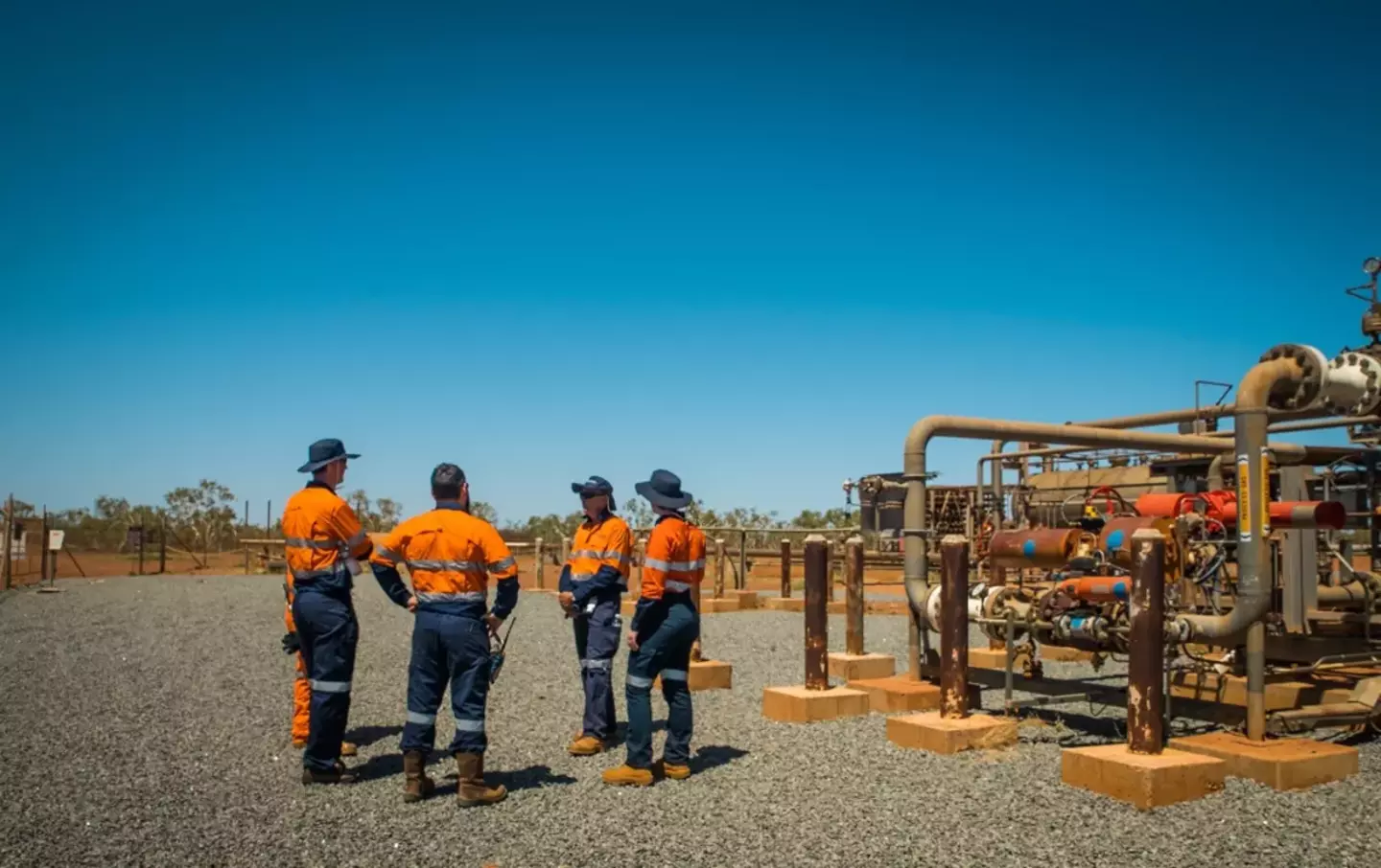 Five workers in high-visibility orange workwear are discussing in front of industrial piping at a sunlit outdoor facility.