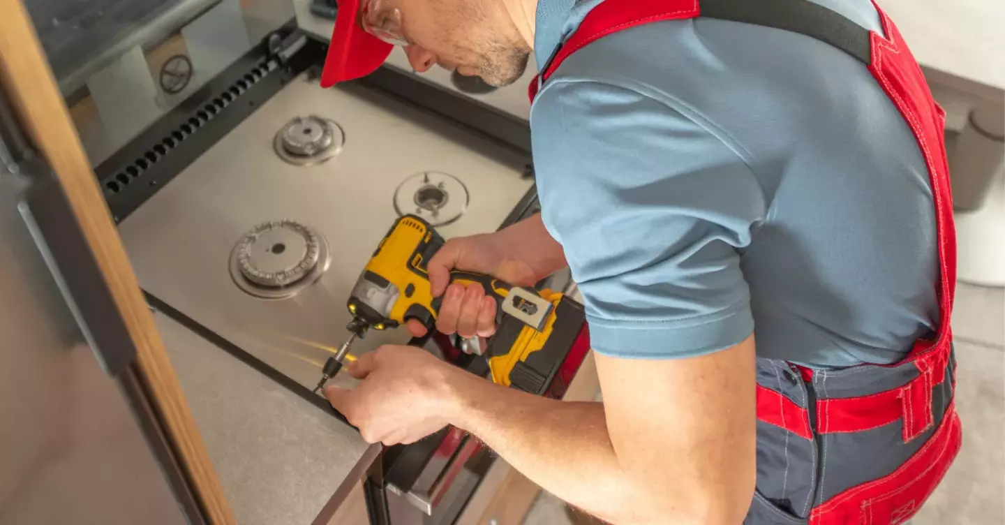 A technician uses a yellow power drill to maintain stove burners in a modern kitchen. The focus is on the maintenance work, underscored by the kitchen environment.