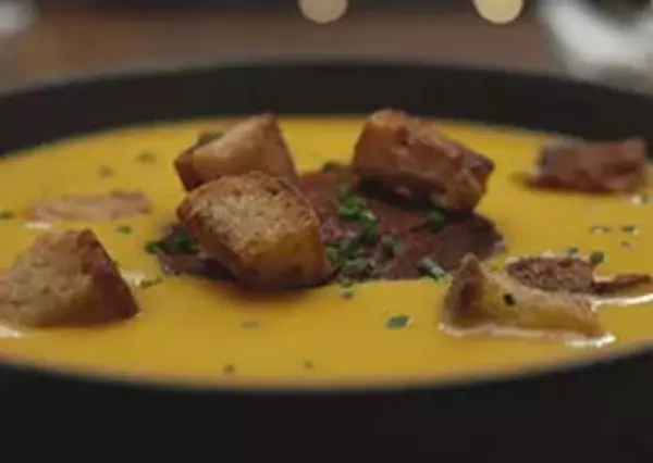 A bowl of creamy soup garnished with croutons and bacon bits, served on a wooden table. The focus is culinary delight in a cozy dining setting.
