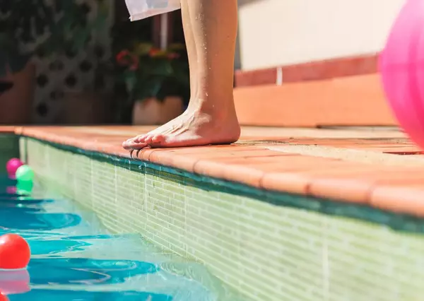 A person stands on the edge of a sunlit swimming pool lined with colorful floating balls, poised to enter the clear turquoise water, suggesting a relaxing, leisurely setting.