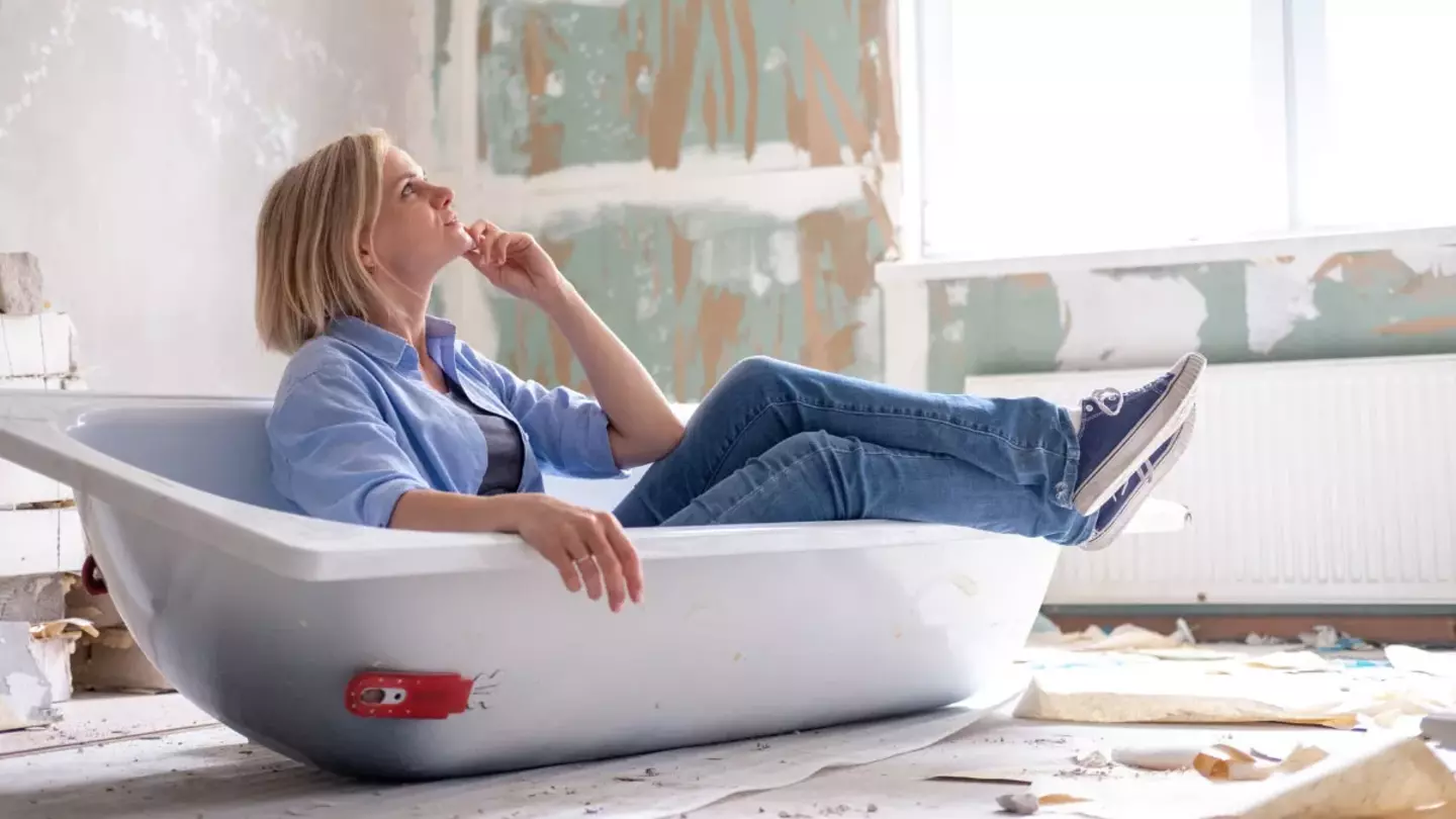 A woman reclines thoughtfully in a bathtub placed in a derelict room with peeling walls and scattered debris, conveying a sense of contemplation amidst renovation.