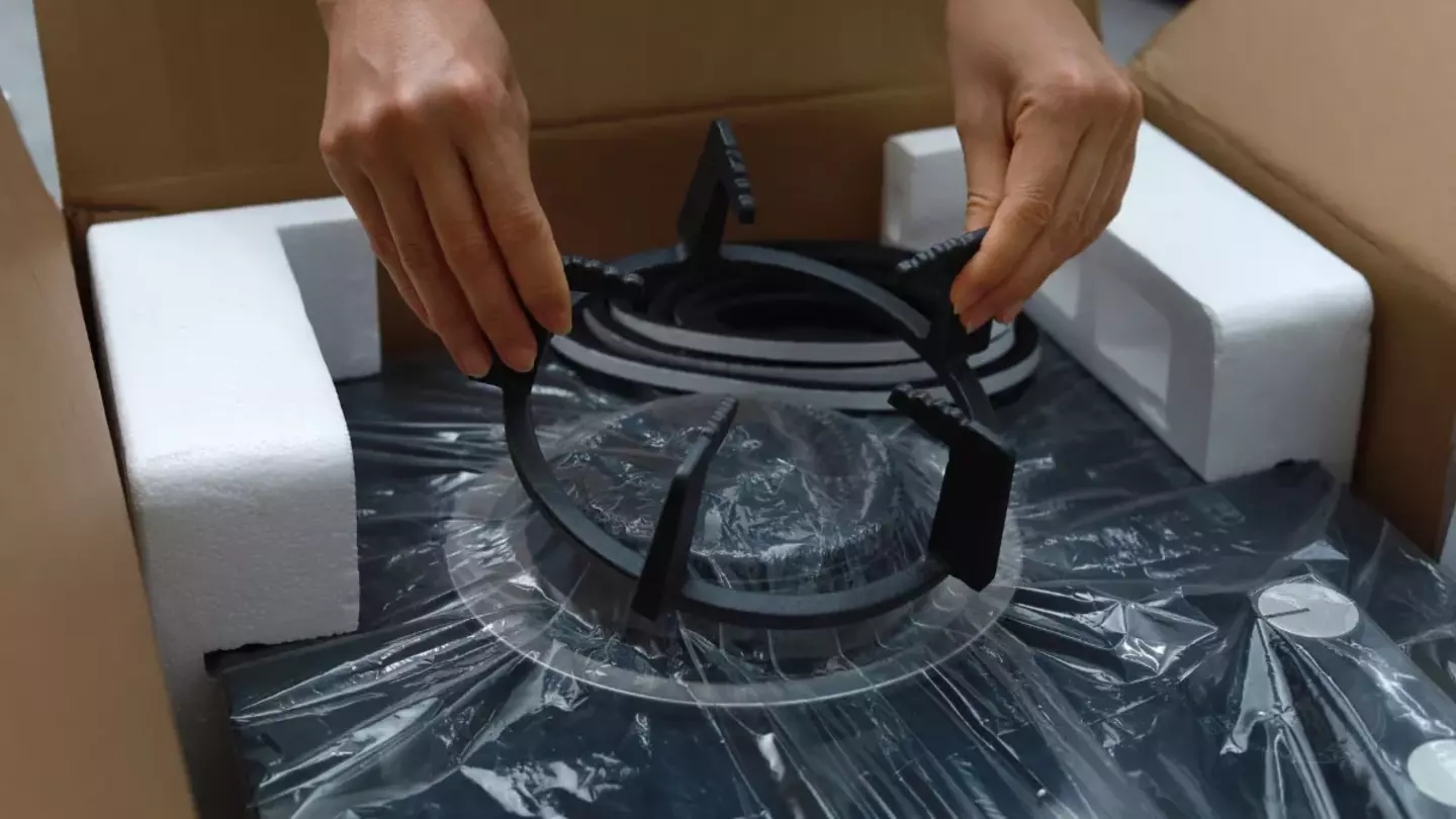 Hands unpack a round, black, coiled stove element from a cardboard box, surrounded by foam padding and plastic wrapping, emphasizing unboxing in a protective packaging environment.