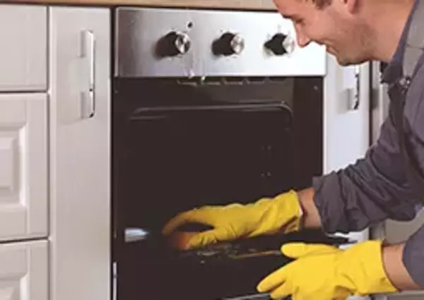 A man wearing yellow gloves cleans an oven, kneeling in a kitchen with white cabinets.