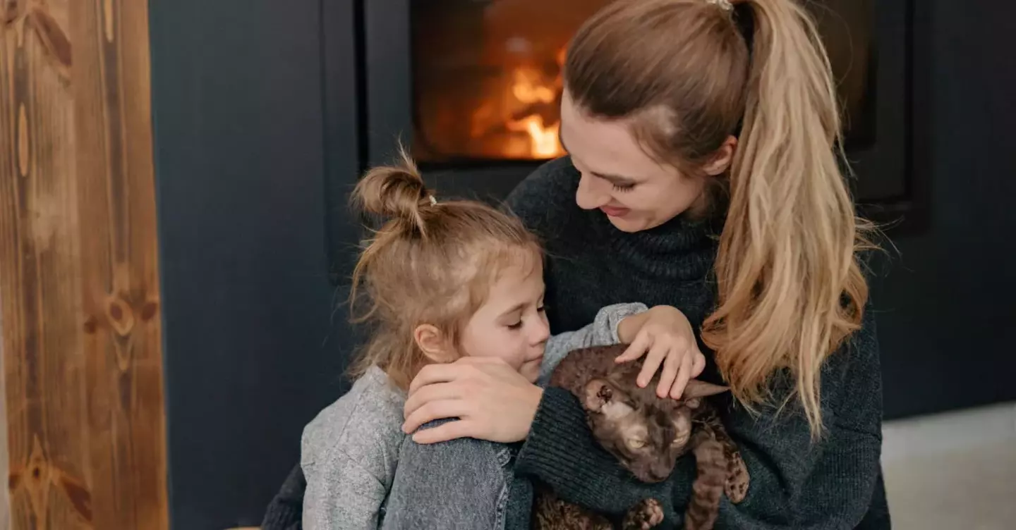 A woman and a young child tenderly embrace a cat near a cozy fireplace. They appear affectionate and calm in a warmly lit, modern living environment.
