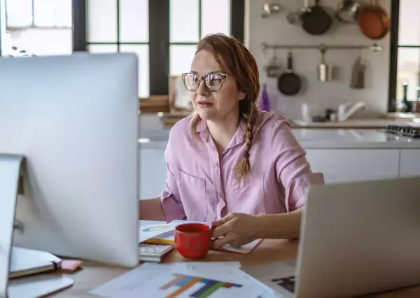 A woman in a pink shirt works intently on a desktop computer in a well-lit, homey kitchen with modern appliances and decor.